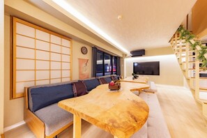 A modern living room with a wooden dining table, blue sofa, and a TV mounted on the wall. - Central Namba and Dotonbori | Spacious 7-bedroom, 1-living room, 1-dining room, 1-kitchen unit with a panoramic open-air bath and BBQ, accommodating up to 14 guests (大阪市)