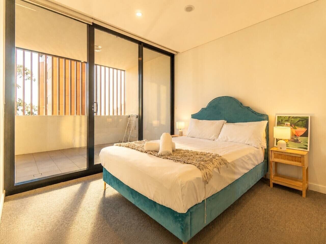 A bedroom with a bed, a nightstand, a lamp, and a view of a balcony.