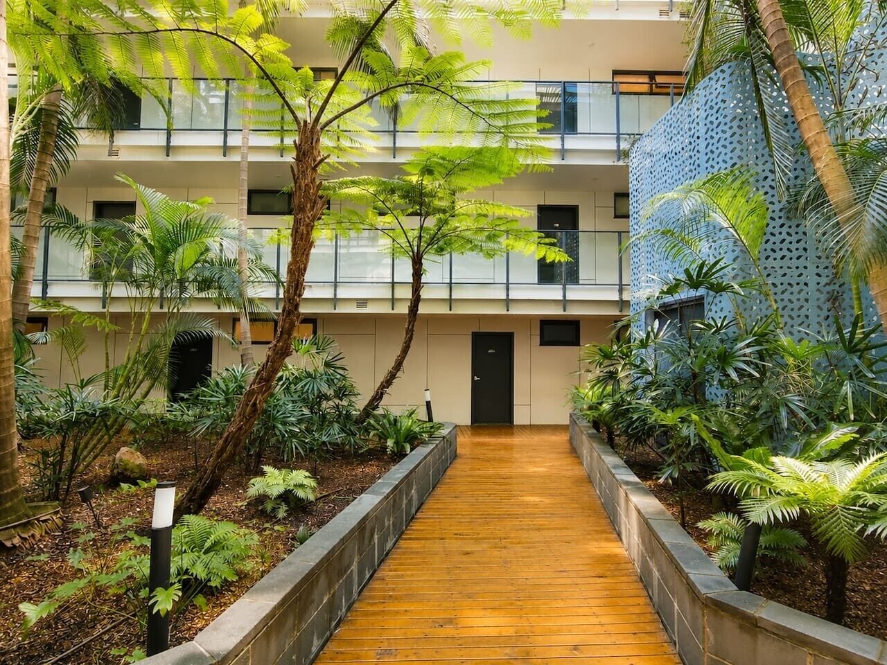 A wooden pathway leading to a building entrance, surrounded by lush greenery and a modern architectural design.
