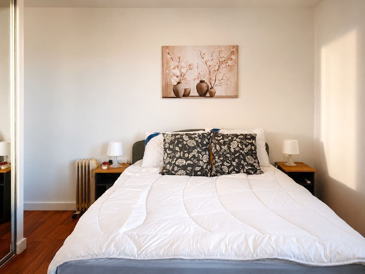 A neatly made bed with white bedding, two bedside tables with lamps, a radiator, and a wall-mounted artwork above the bed.