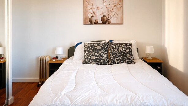 A neatly made bed with white bedding, two bedside tables with lamps, a radiator, and a wall-mounted artwork above the bed. - Dee Why 2BR Apartment Near Beach & Shops (Dee Why)