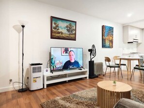 Ein Wohnzimmer mit Flachbildfernseher, einer weißen Unterhaltungseinheit, einem Standventilator und einem Essbereich mit Stühlen und Tisch.