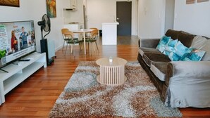 A living room with a TV, sofa, coffee table, and a dining area with chairs and a table. - Dee Why 2BR Apartment Near Beach & Shops (Dee Why)
