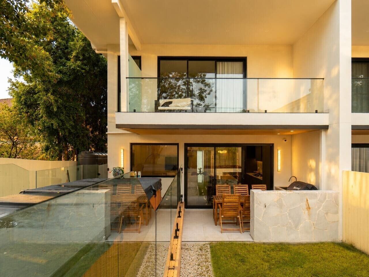 A modern two-story house with a glass-enclosed patio, outdoor seating, and a barbecue area.