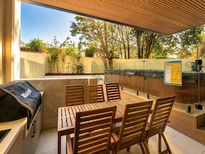 An outdoor patio with a wooden table and chairs, a barbecue grill, and a glass enclosure.