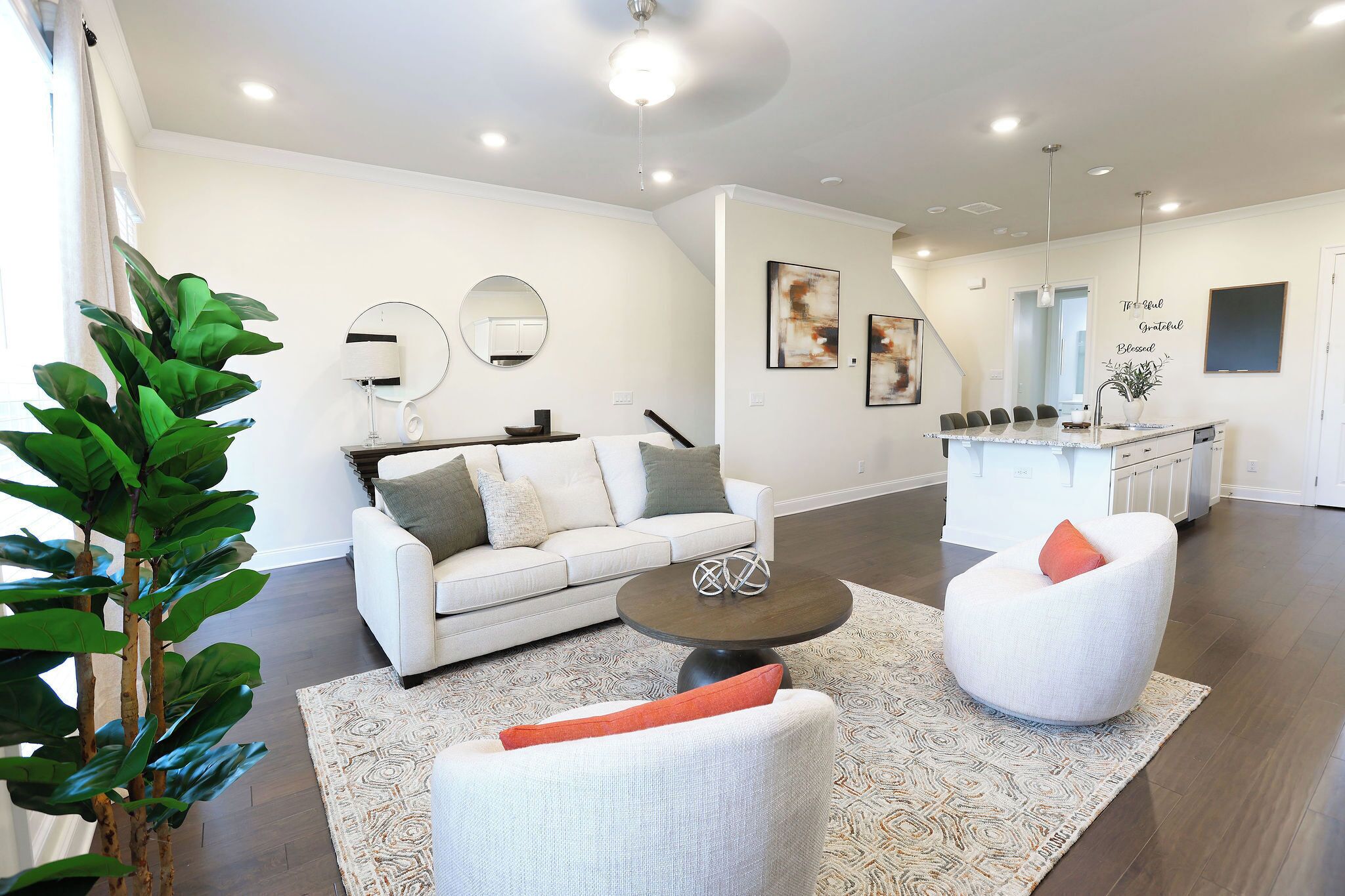 A modern living room with a white sofa, a round coffee table, and a white armchair with orange cushions. There is a large plant in the corner, a dining area with a kitchen in the background, and artwork on the walls.