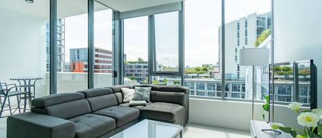En moderne stue med en hjørnesofa, et glas sofabord og store vinduer med udsigt over byen.