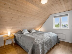 Une chambre avec un plafond en pente, un lit recouvert d’une couverture à motifs, deux lampes de chevet et une fenêtre donnant sur un paysage verdoyant.