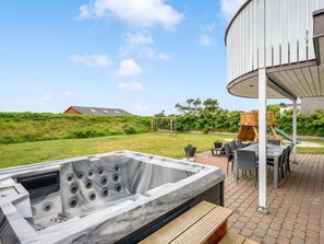 Ein Whirlpool mit einer hölzernen Sitzbank, eine überdachte Terrasse mit Gartenmöbeln und Blick auf ein Wiesenfeld sowie ein Gebäude im Hintergrund.