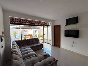 Uma sala de estar ampla com um sofá seccional grande, uma TV de tela plana na parede e uma porta de vidro deslizante para uma área externa.