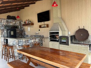 A bar area with a wooden bar counter, stools, a television mounted on the wall, and a brick oven. - Recanto do Benji - Piscina e Sauna- Proximo a praia (Santa Cruz Cabrália)