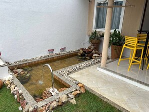 A small outdoor water feature with a fountain, surrounded by rocks and grass, with a yellow chair and a table nearby.
