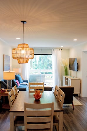 A modern living room with a dining table, chairs, a sofa, a TV, and a large window with a view of trees. - Coastal Oasis Near Coligny Beach (Hilton Head Island)