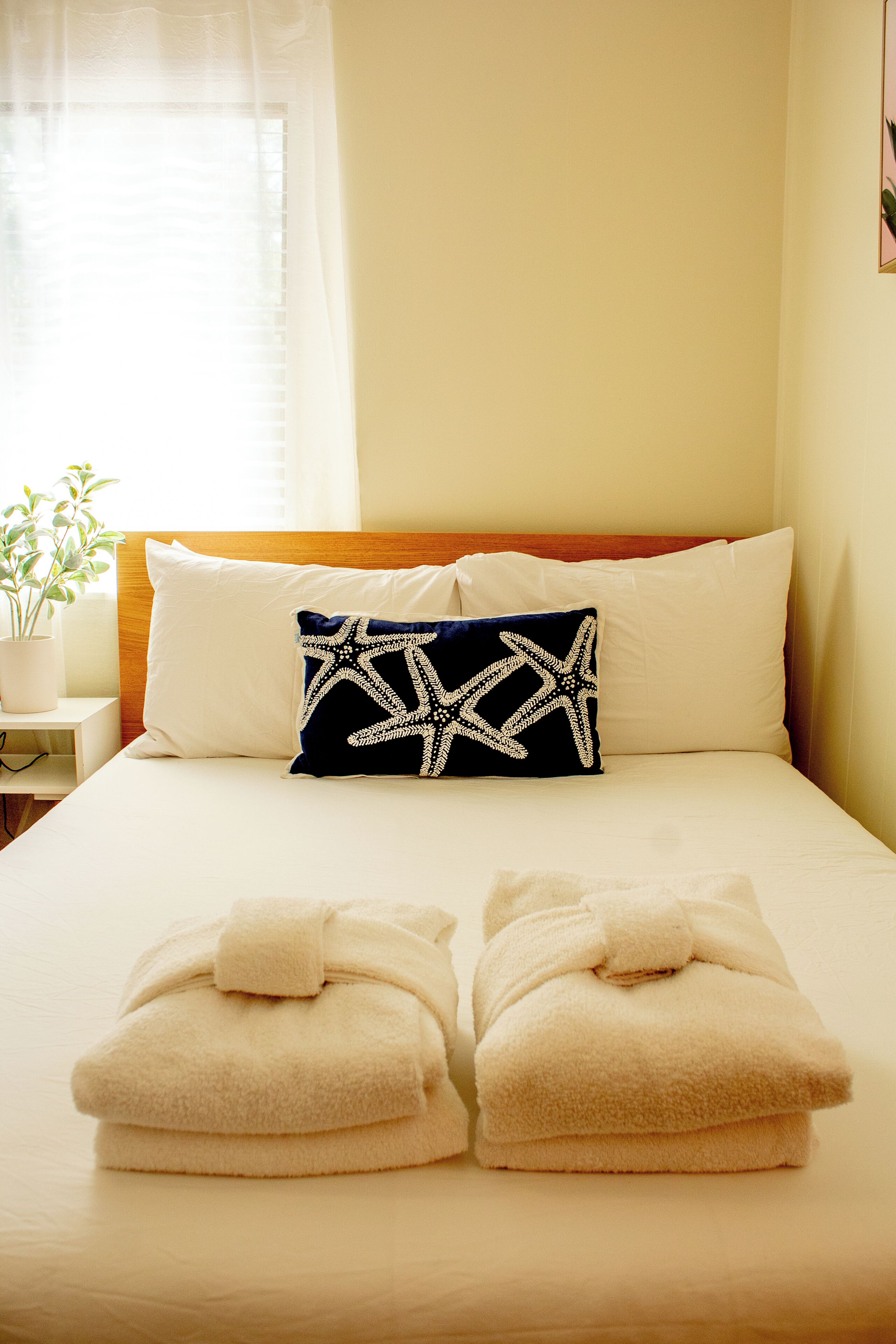Un lit bien fait, avec du linge de lit blanc, une tête de lit en bois et un coussin décoratif orné d’un motif en forme d’étoile de mer.