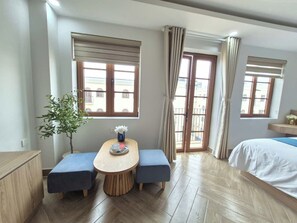 Ein Hotelzimmer mit Holzboden, einem Bett, einem Esstisch mit zwei blauen Bänken, einer Topfpflanze und Blick durch die Glastüren.