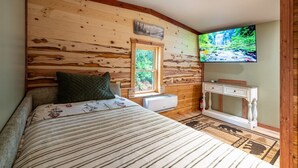 A bedroom with a wooden wall, a bed with a floral bedspread, a window, a white bedside table, and a television on the wall. - Firefly Retreat - Upscale Off-Grid Mountain Cabin (Marble Hill)