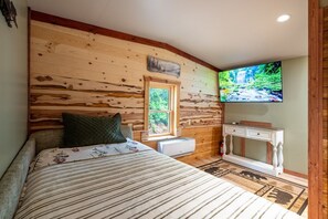 Ein Schlafzimmer mit Holzwand, einem Bett mit floraler Bettdecke, einem Fenster, einem weißen Nachttisch und einem an der Wand befestigten Fernseher.