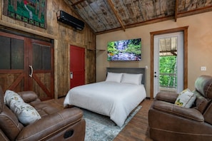Ein Schlafzimmer mit einem Bett, zwei Ledersesseln, einer roten Tür, einem an der Wand befestigten Fernseher und Blick auf Grün durch ein Fenster.