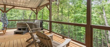 A covered outdoor deck with wooden flooring, metal chairs, a barbecue grill, and a view of a forested area.