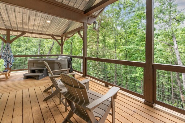 A covered outdoor deck with wooden flooring, metal chairs, a barbecue grill, and a view of a forested area.