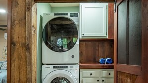 A compact laundry room with a washing machine and dryer, wooden cabinets, and a small shelf with folded towels. - Firefly Retreat - Upscale Off-Grid Mountain Cabin (Marble Hill)