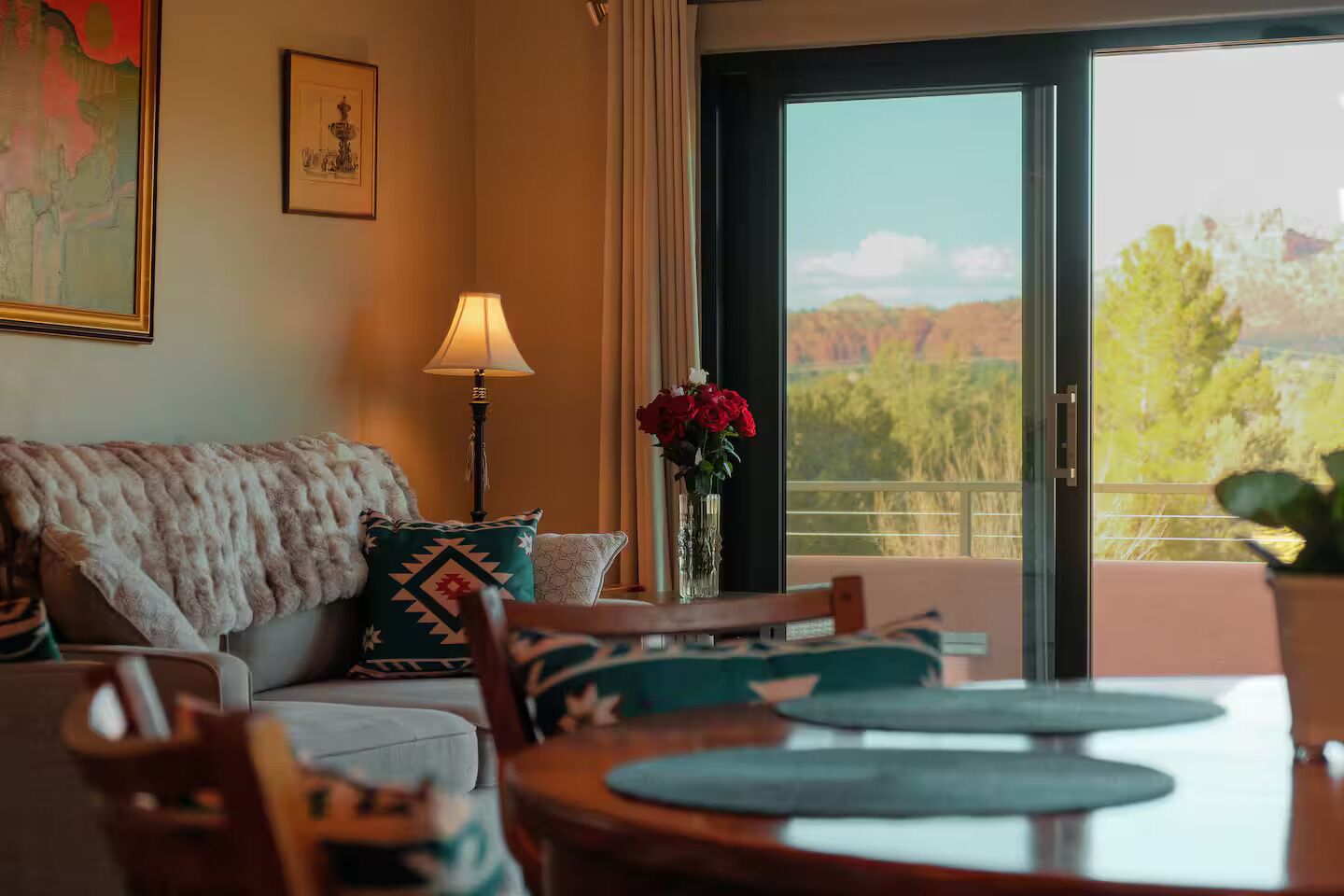 A room with a sofa, a table with chairs, a lamp, a vase with flowers, and a view through a glass door.