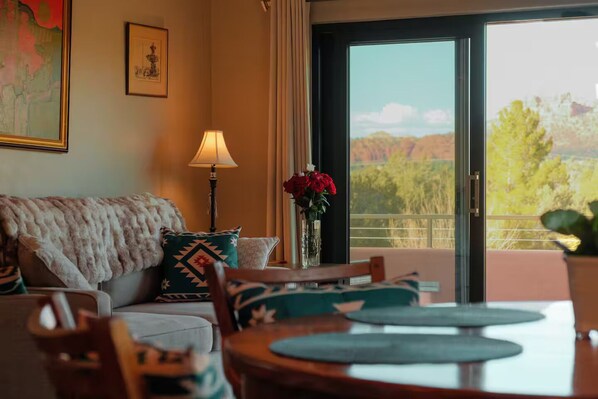 Una habitación con sofá, una mesa con sillas, una lámpara, un florero con flores y vistas a través de un ventanal.