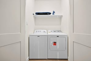 A laundry room with two washing machines and a dryer, a shelf above the machines, and a door with a safety lock. - Blue Lagoon • New Villa Near Beach (Galveston)