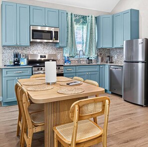 A kitchen with teal cabinets, stainless steel appliances, a wooden dining table with chairs, and a window with curtains. - Blue Lagoon • New Luxury Beach Villa  • Sleeps 5 (Galveston)