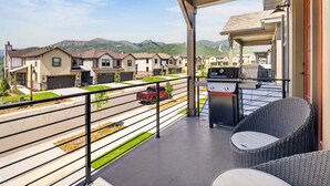 A covered balcony with a grill and wicker furniture, overlooking a street with houses and mountains. - Mountain Haus, Hot Tub, Sauna, Deer Valley Close (Heber City)