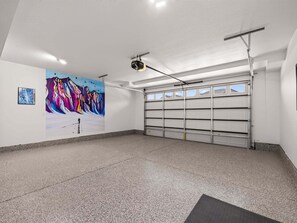 A garage with a large, colorful abstract painting on the wall, a ceiling-mounted projector, and a garage door with multiple storage compartments. - Mountain Haus, Hot Tub, Sauna, Deer Valley Close (Heber City)
