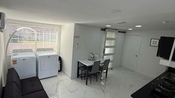 A compact living space with a sofa, a dining table with chairs, a washing machine, and a small kitchen area with a stove.