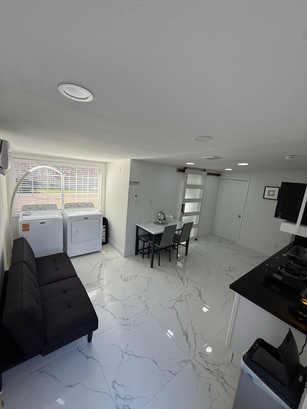 A compact living space with a sofa, a dining table with chairs, a washing machine, and a small kitchen area with a stove.