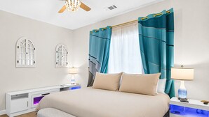 Quarto moderno com cama, mesas de cabeceira, escrivaninha, ventilador de teto e janela com cortinas.