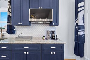 A kitchen with dark blue cabinets, a stainless steel microwave, a coffee maker, and a tiled backsplash. - Twilight Harbor • New Villa Near Beach (Galveston)