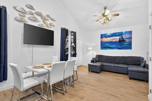 A modern living room with a flat-screen TV, a gray sofa, a dining area with white chairs, and a ceiling fan. - Twilight Harbor • New Luxury Beach Villa•Sleeps 5 (Galveston)