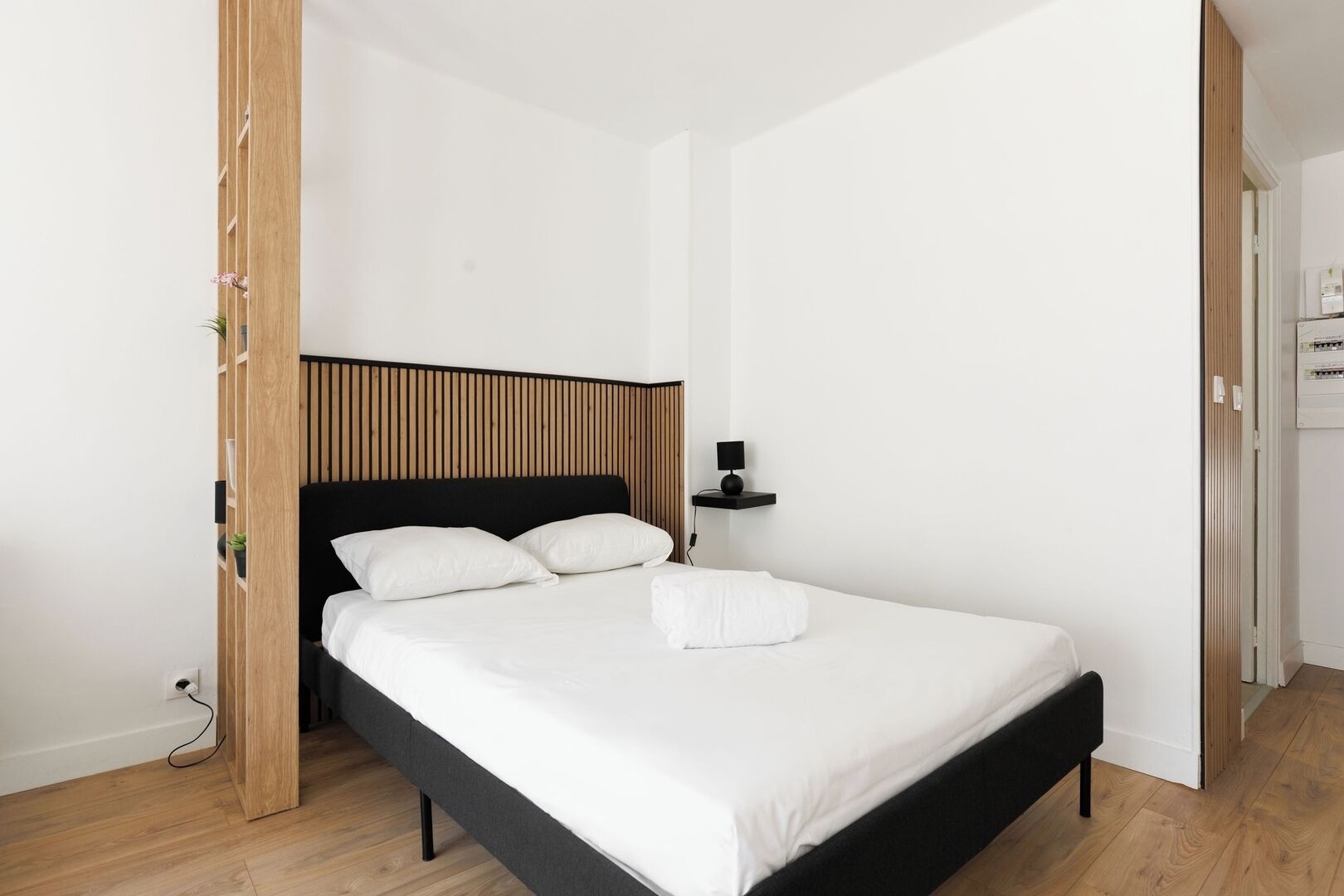 Un letto singolo con struttura nera e biancheria bianca, un comodino con una lampada e un pannello di legno su un lato.