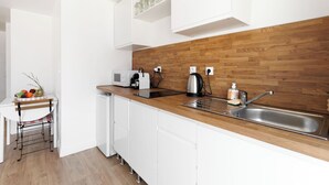 Uma cozinha compacta com armários brancos, bancada de madeira, pia, chaleira, máquina de café e uma pequena área de refeições com uma cadeira e mesa.
