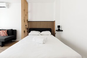 A modern hotel room with a large bed, a wooden headboard, a bedside table with a lamp, and a dark-colored sofa with a patterned pillow. - Upstays Studio 28m2 Balcony AC (Cannes)