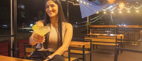 A person seated at a wooden table with a drink, under a canopy with string lights.