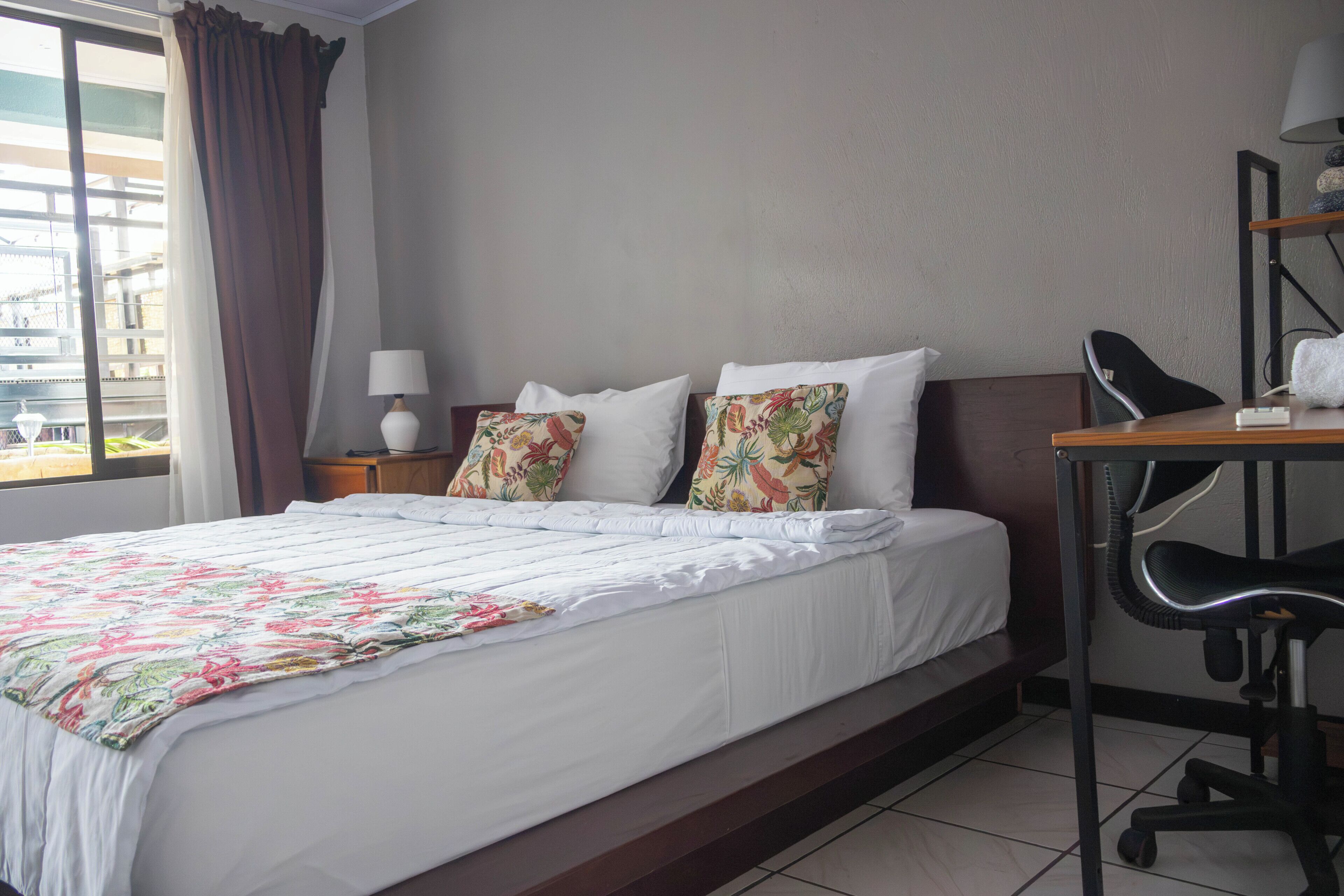Habitación de hotel con una cama grande, un escritorio con silla, una lámpara y un ventanal con cortinas.