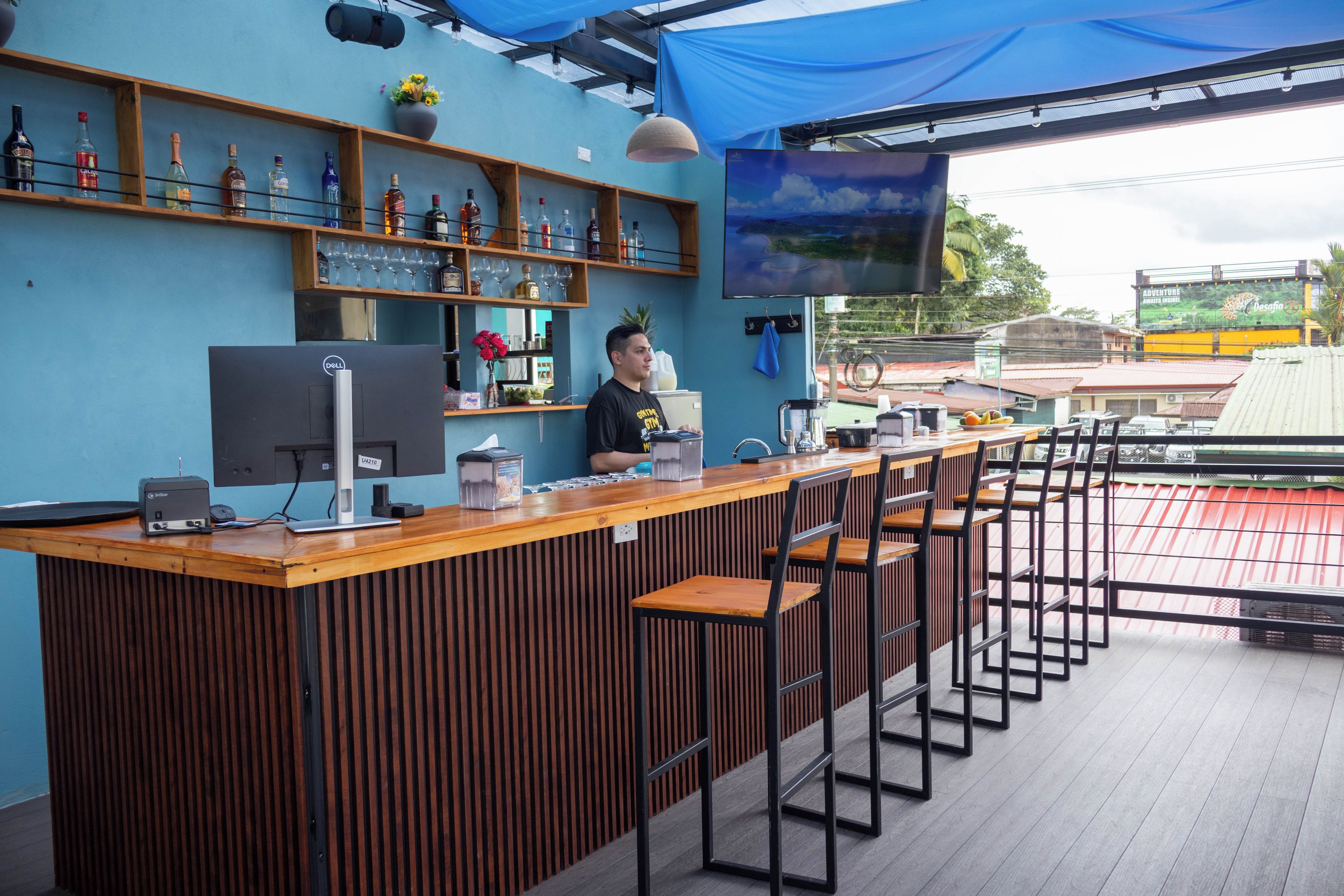 Uma área de bar com balcão de madeira, banquetas, um computador e prateleiras com garrafas e copos.