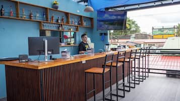 Uma área de bar com balcão de madeira, banquetas, um computador e prateleiras com garrafas e copos.