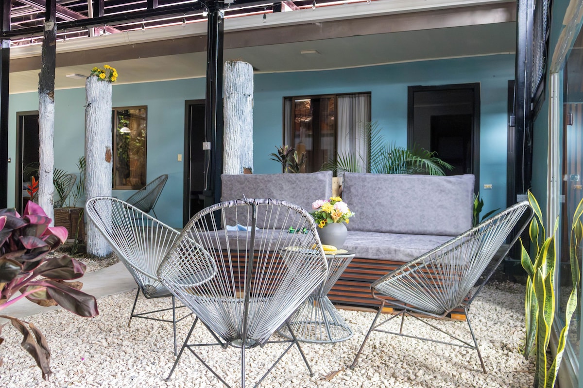 Un área de estar moderna al aire libre con sofá y sillas de estructura metálica, rodeada de plantas y un suelo de grava.