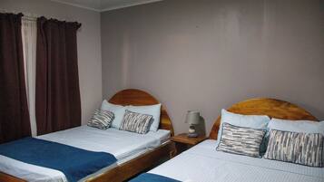 Habitación de hotel con dos camas individuales, cada una con cabecera de madera, una mesita de noche con lámpara y una ventana con cortinas.