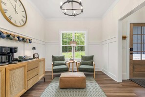 A room with a large wall clock, a coffee machine, two armchairs, a small table, and a window with a view of greenery.