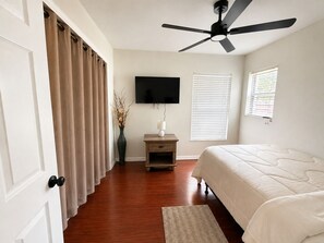 Quarto com cama, ventoinha no teto, televisão, mesa de cabeceira, um vaso com flores e janela com persianas.