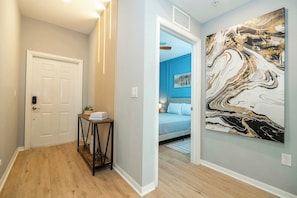 Un pasillo con una puerta, una mesita con libros, una pared con un cuadro abstracto grande y un dormitorio con una cama y una mesita de noche.