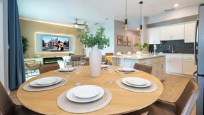 Un moderno comedor con una mesa redonda dispuesta para seis personas, un televisor mostrando una escena de castillo y una cocina al fondo.