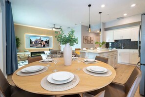 A modern dining area with a round table set for six, a television displaying a castle scene, and a kitchen in the background. - Cozy 3BR Apartment Storey Lake Resort Near Disney (Kissimmee)